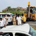 Police officers and bystanders at the scene of a crushed car accident with an ambulance and excavator in the background