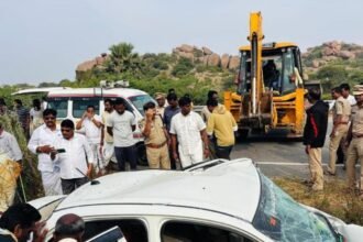 Police officers and bystanders at the scene of a crushed car accident with an ambulance and excavator in the background