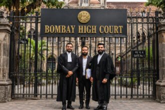 Two lawyers in black robes standing with their client in front of the Bombay High Court gate