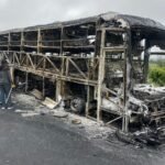 urnt-out skeleton of a bus on a roadside with officials standing nearby