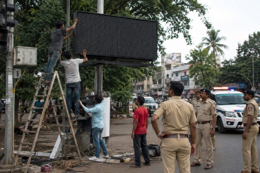 णे महापालिका कर्मचारी शिडीवर चढून रस्त्यावरील डिजिटल फलक काढताना, पार्श्वभूमीत पोलीस व पोलीस वाहन उभे