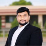 ॲड. विराज करचे यांचा औपचारिक पोर्ट्रेट फोटो, काळ्या ब्लेझरमध्ये, इमारतीसमोर उभे