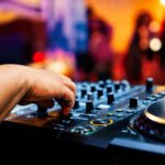 A DJ's hand adjusting a mixing console at a nighttime event with a crowd in the background