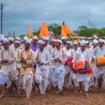पांढरा पोशाख व भगवे फेटे परिधान केलेले वारकरी ढोल वाजवत मिरवणुकीत चालताना