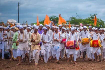 पांढरा पोशाख व भगवे फेटे परिधान केलेले वारकरी ढोल वाजवत मिरवणुकीत चालताना